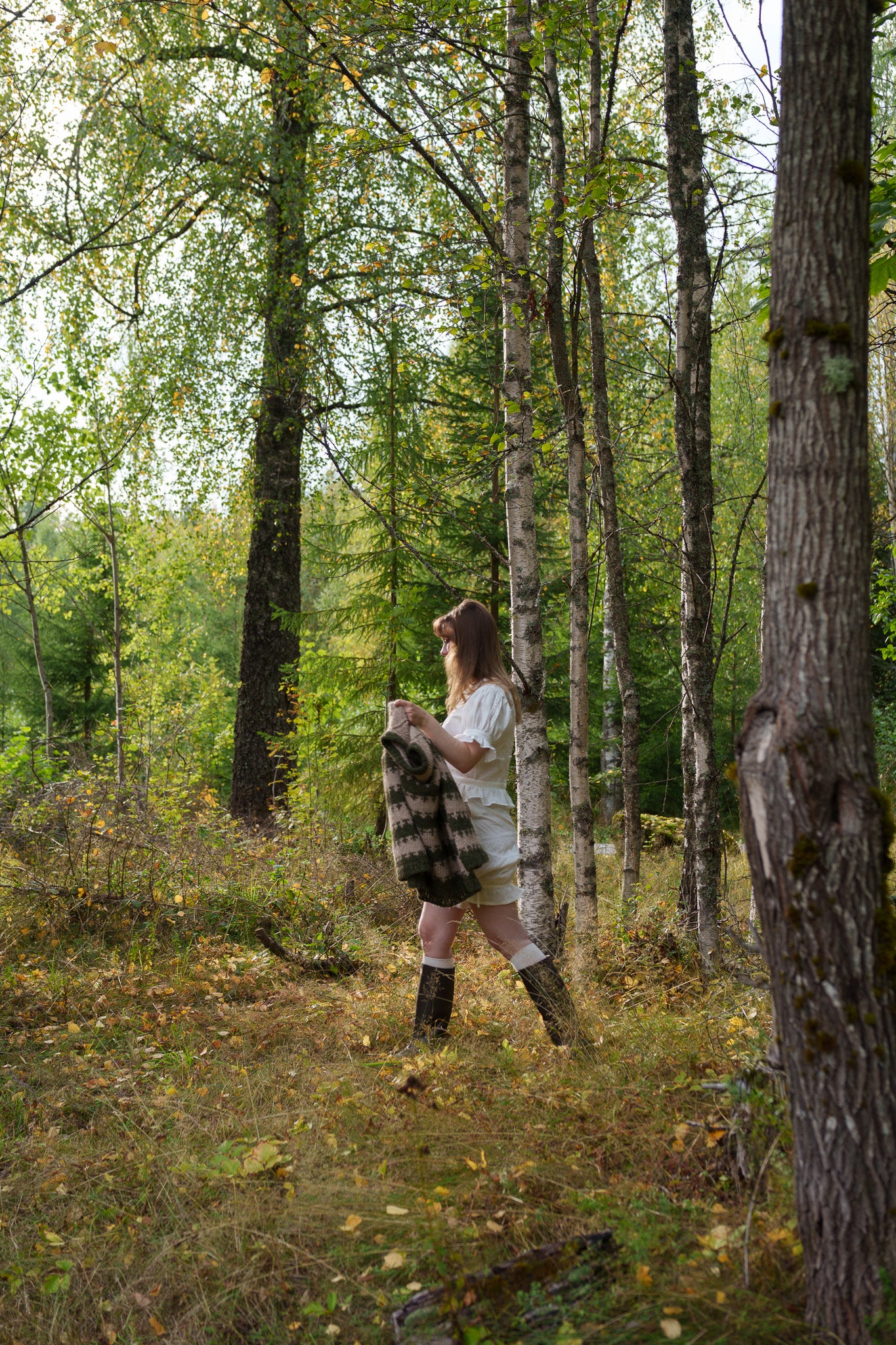 Person standing in a forest holding a sweater, surrounded by trees and foliage.