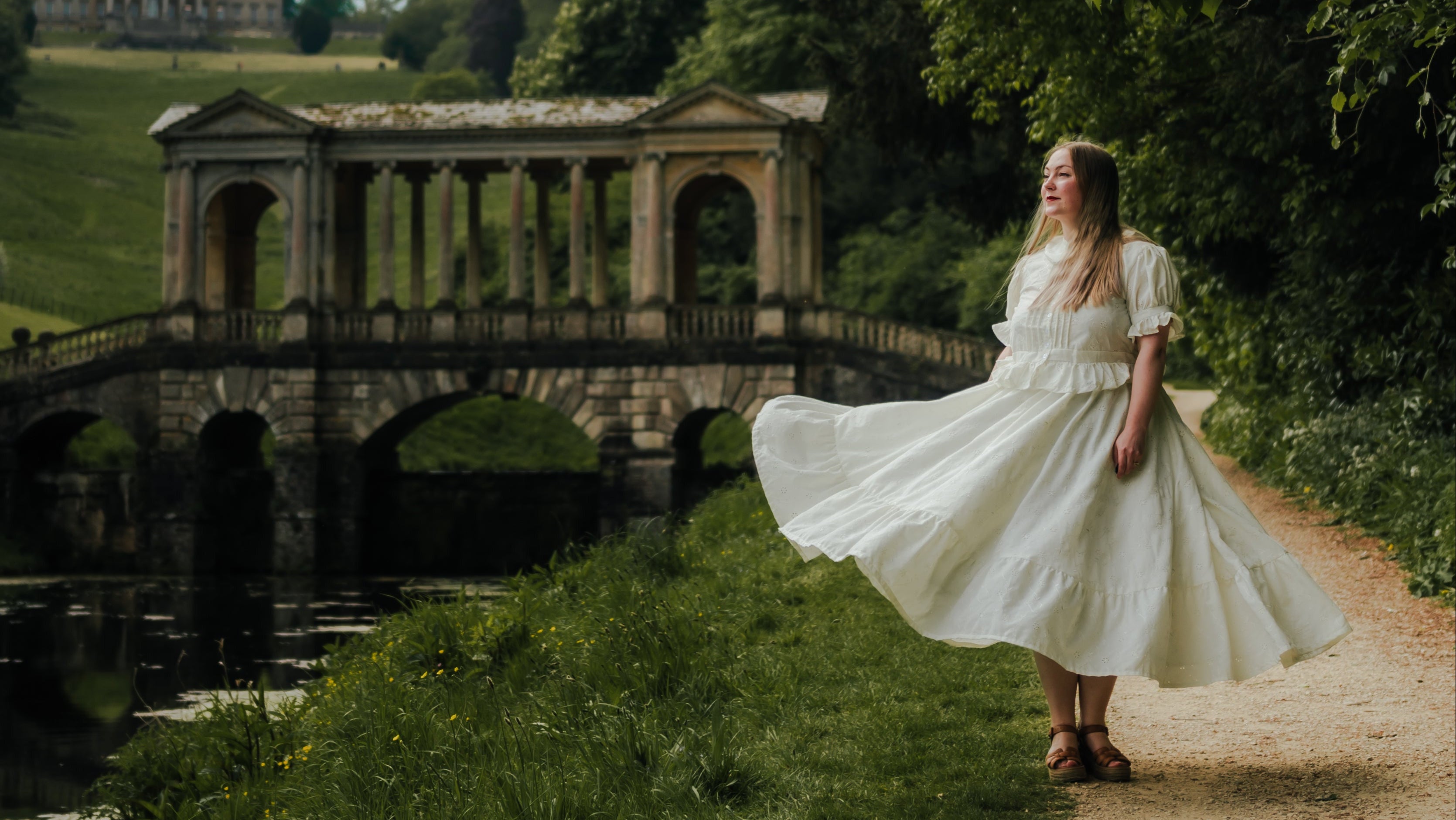 A woman in a flowing white dress stands on a dirt path beside a canal, with a stone bridge and green parkland in the background under a cloudy sky.