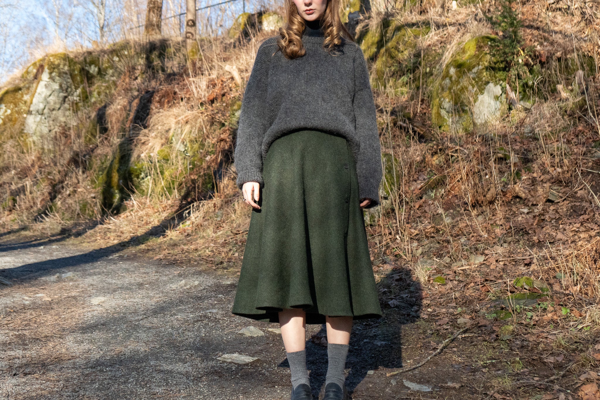 Person wearing a dark gray sweater and green skirt standing on a path with a natural background