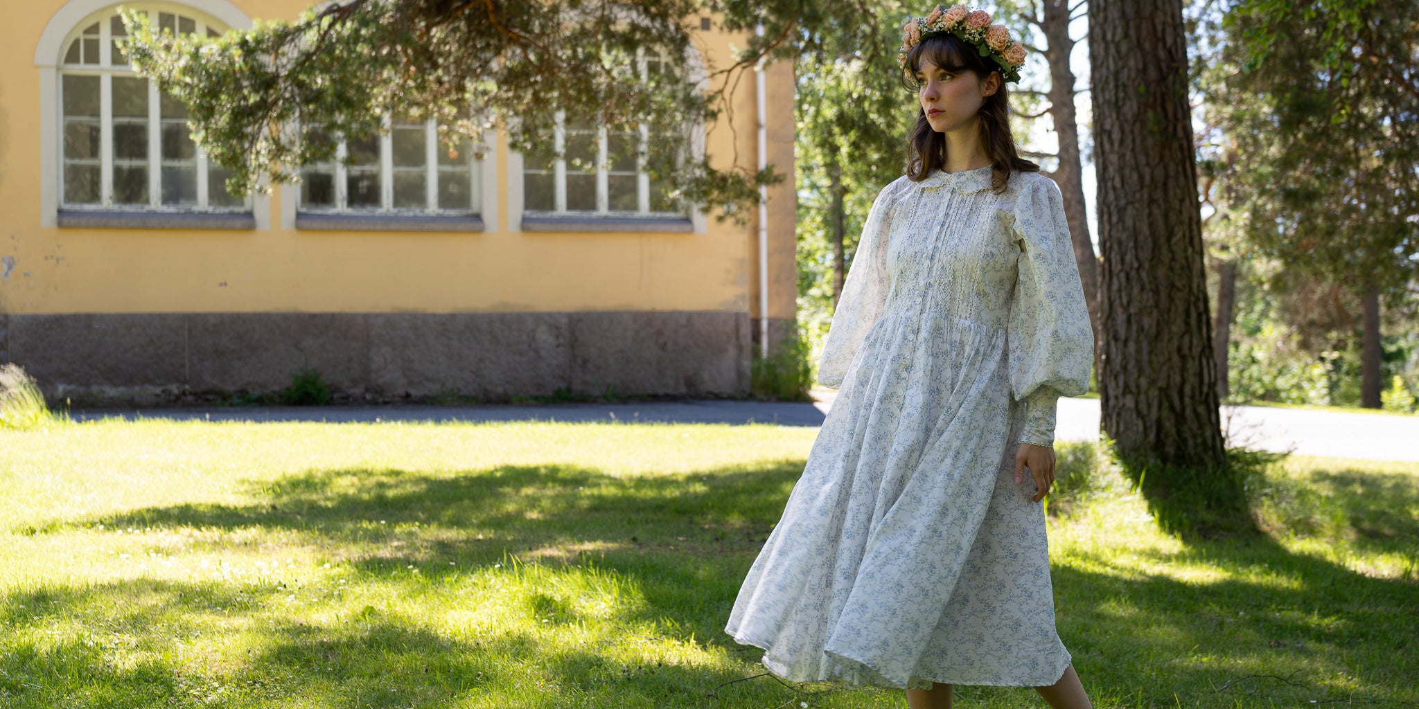A woman wearing a long white lace dress stands outdoors on a grassy lawn beside a tree, with a yellow building in the background.