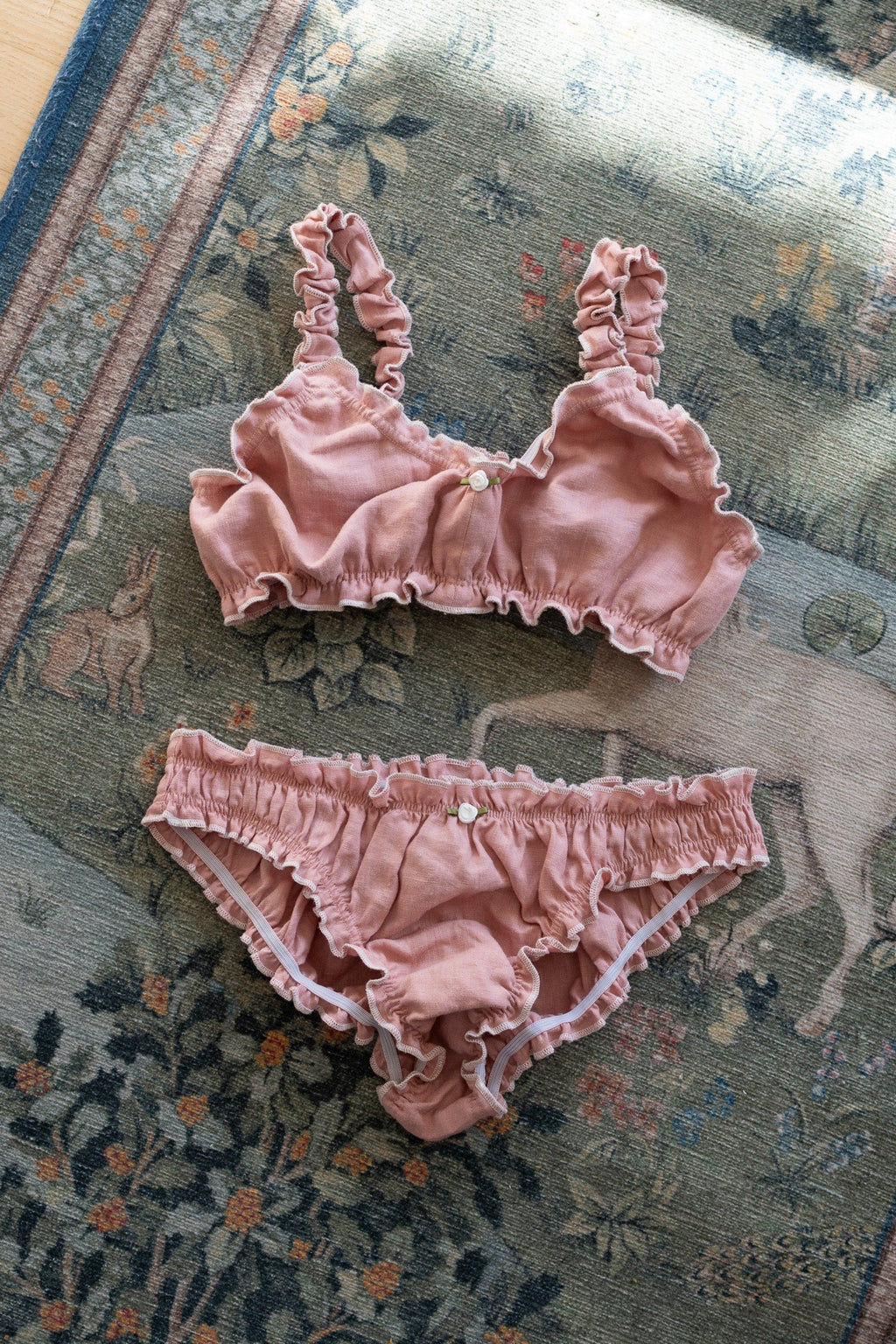 A two-piece baby lingerie set in dusty rose pink ruffled fabric, consisting of a bralette with side buttons and matching briefs, laid flat on a patterned rug.