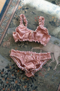 A two-piece baby lingerie set in dusty rose pink ruffled fabric, consisting of a bralette with side buttons and matching briefs, laid flat on a patterned rug.
