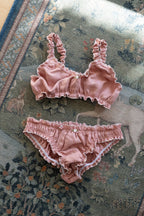 A two-piece baby lingerie set in dusty rose pink ruffled fabric, consisting of a bralette with side buttons and matching briefs, laid flat on a patterned rug.