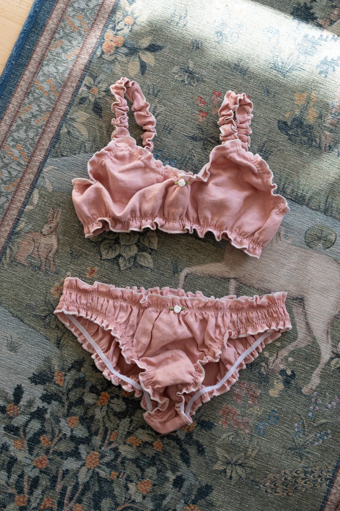A two-piece baby lingerie set in dusty rose pink ruffled fabric, consisting of a bralette with side buttons and matching briefs, laid flat on a patterned rug.