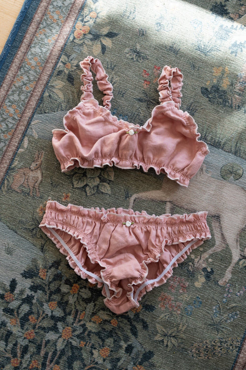 A two-piece baby lingerie set in dusty rose pink ruffled fabric, consisting of a bralette with side buttons and matching briefs, laid flat on a patterned rug.